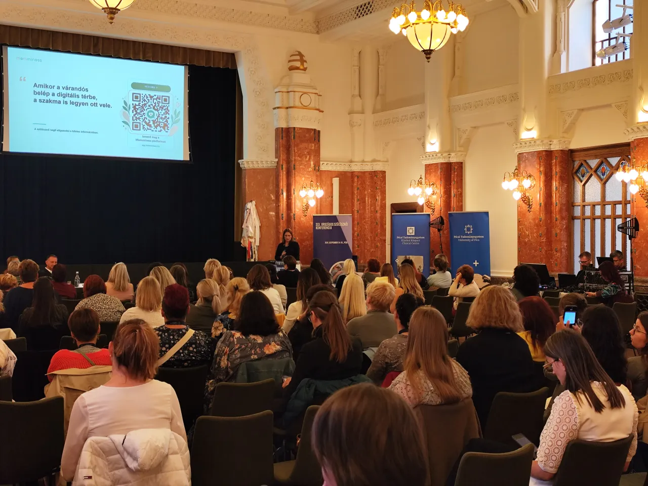 Zsebe-Vasvári Viktória a színpadon tart Momminess-előadást, miközben a konferenciaterem közönsége figyeli.